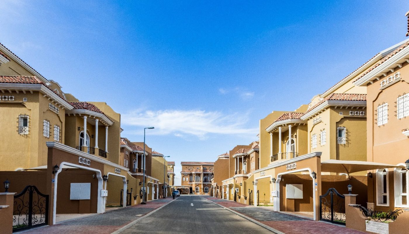 A luxury residential street in Janabiya features modern, multi-story villas in shades of yellow and orange under a clear blue sky. The road is flanked by well-maintained sidewalks and lampposts.