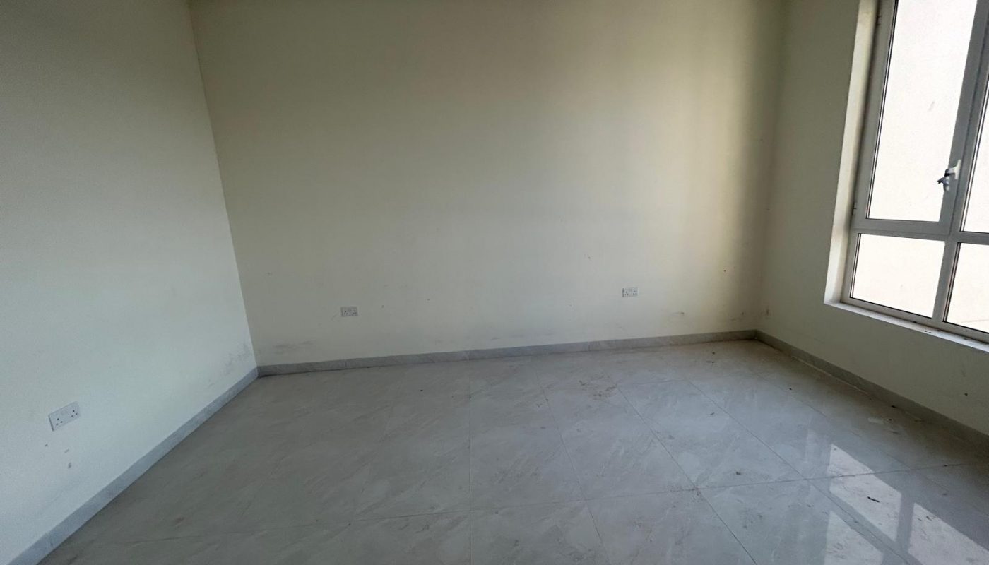 An empty, unfurnished room with cream-colored walls, tiled floor, and a large window letting in natural light.
