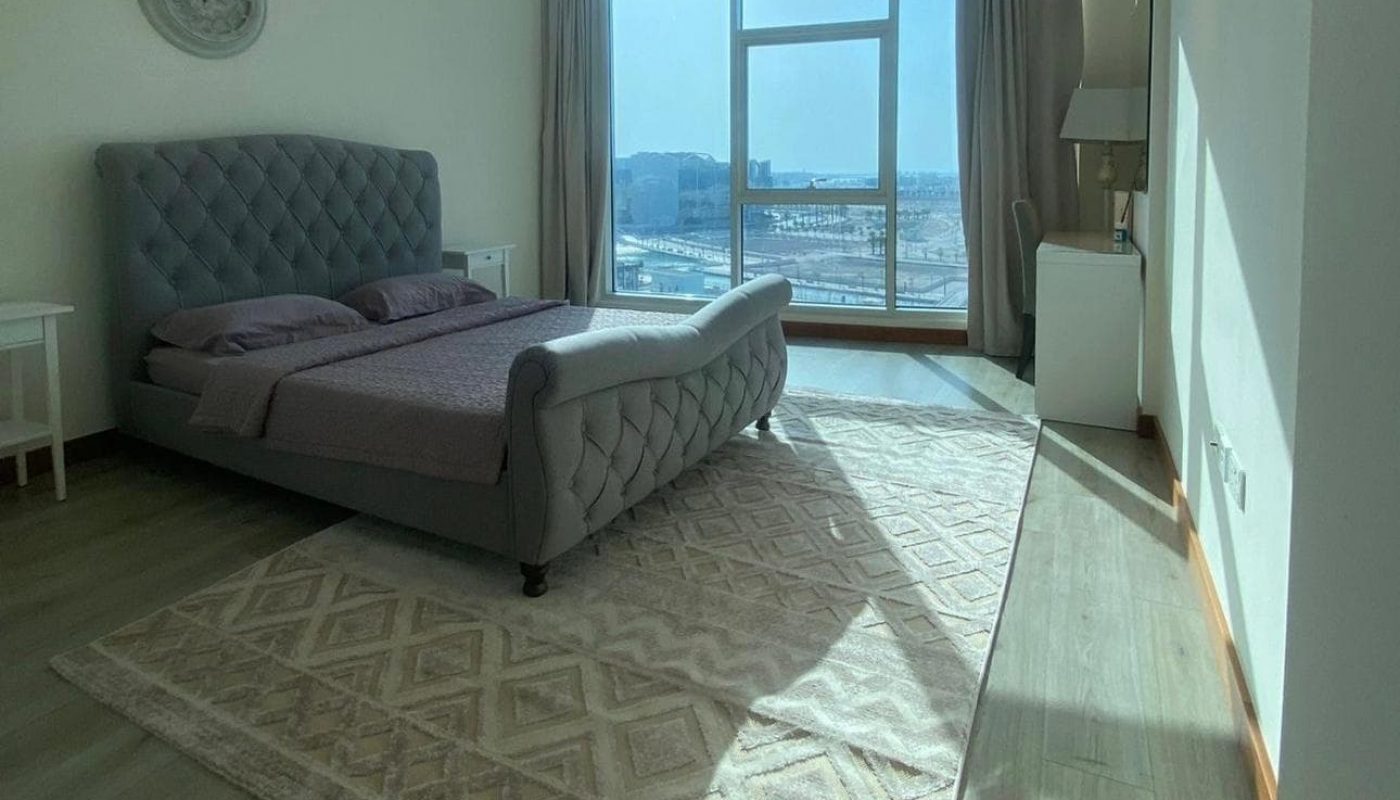 A bright bedroom with a large bed, patterned rug, window with city view, a wall clock, and a desk with a chair.