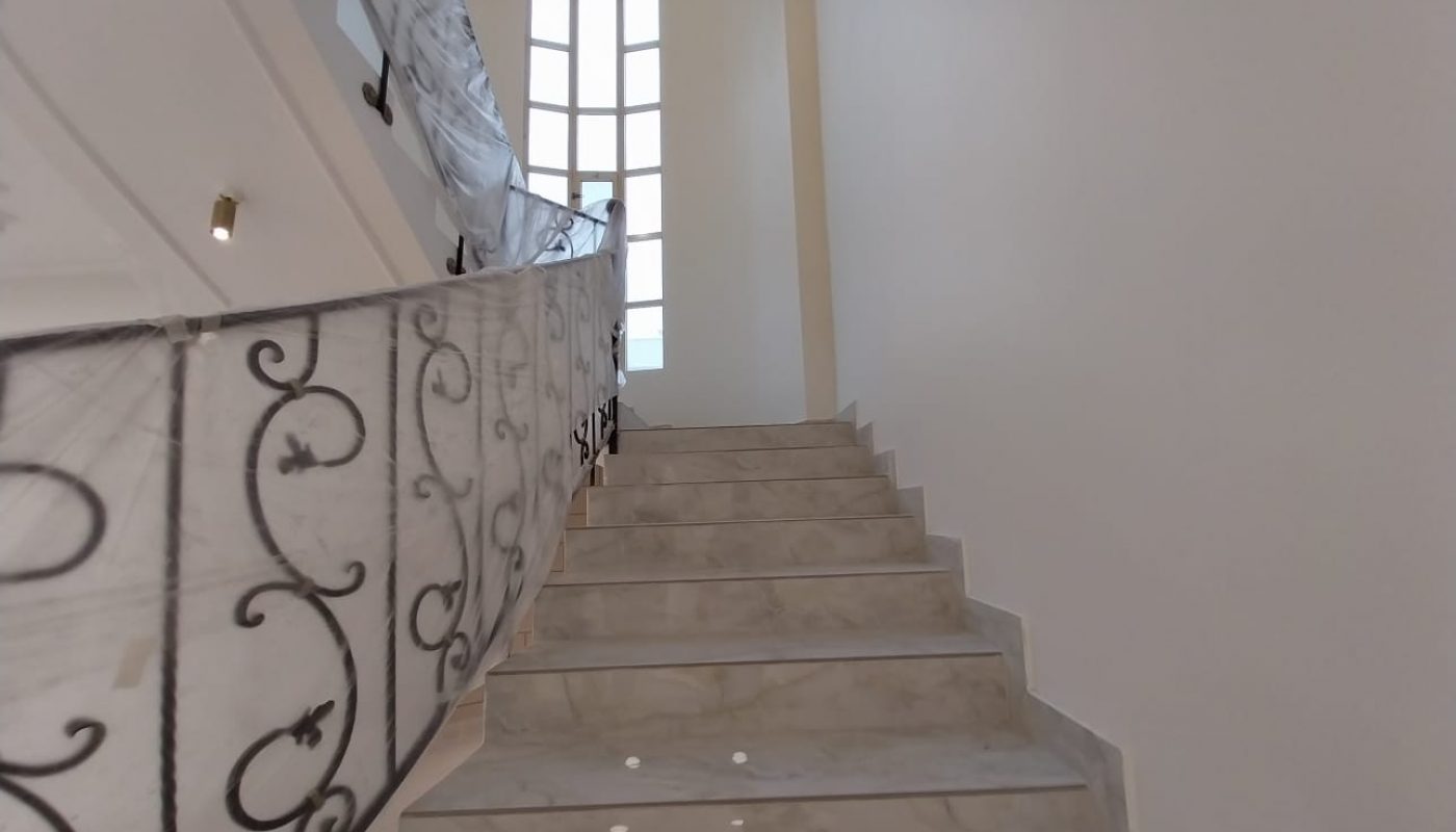 A marble staircase with a decorative black metal railing leads up to a tall, arched window in a white-walled interior space.