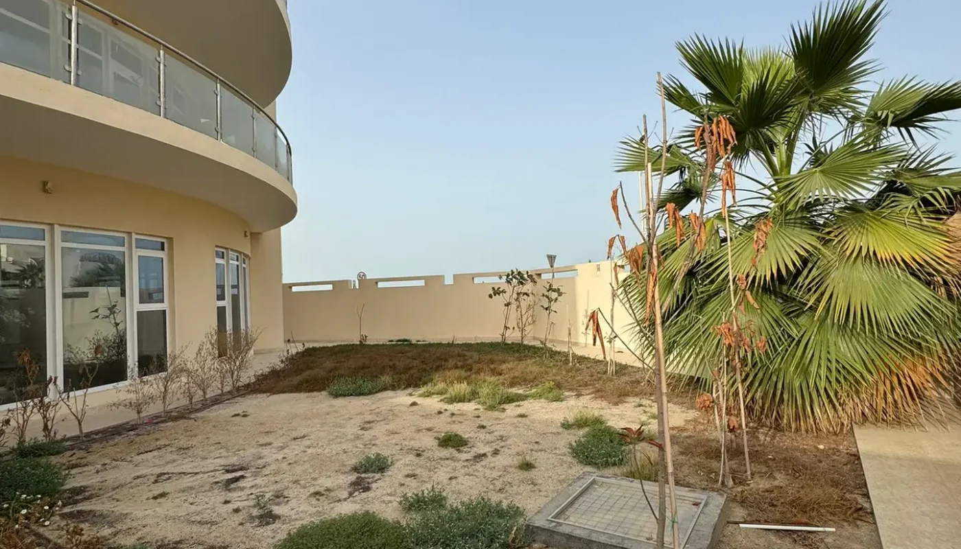 A sandy courtyard with sparse greenery and a palm tree, bordered by a curved building with balconies and a white fence under a clear sky.