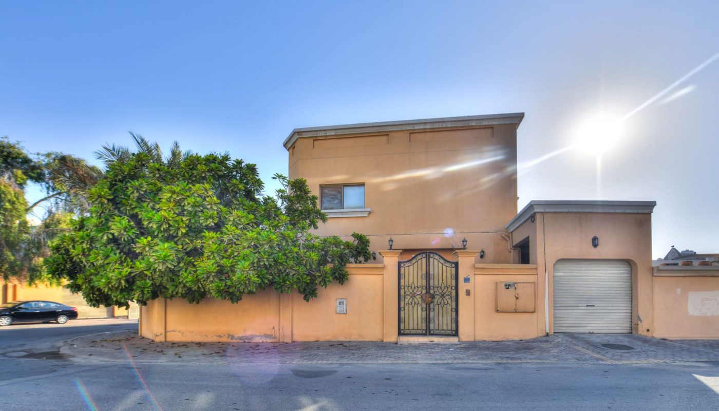 Two-story beige house with a gated entrance, garage door, and large tree in front, situated on a corner under a clear sky with the sun shining.