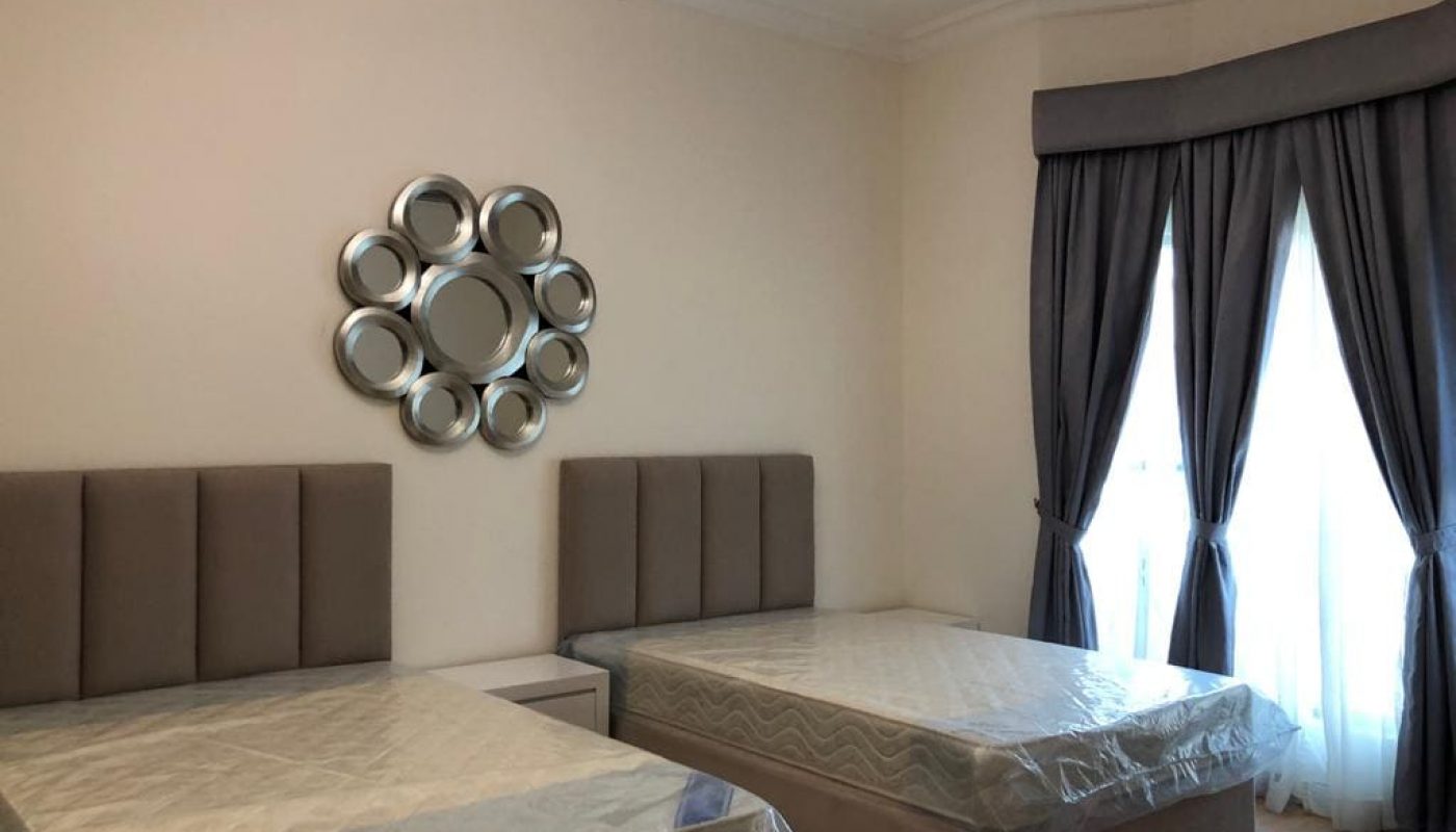 A luxury villa bedroom with two single beds covered in plastic, a decorative circular mirror on the wall, and long gray curtains.