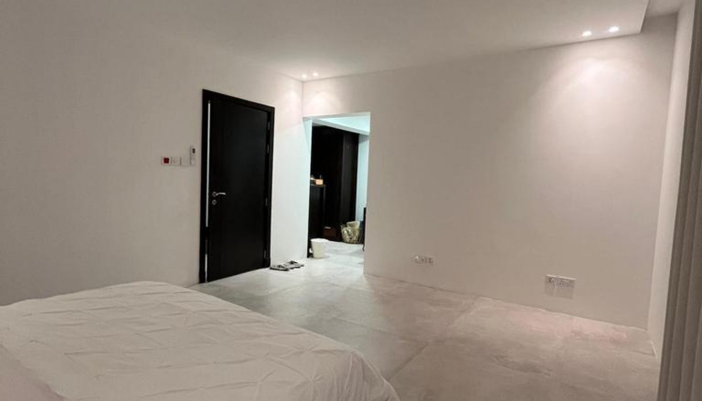 A minimalist bedroom with a white bed, tiled floor, white walls, and black doors. The room is well-lit with recessed ceiling lights.
