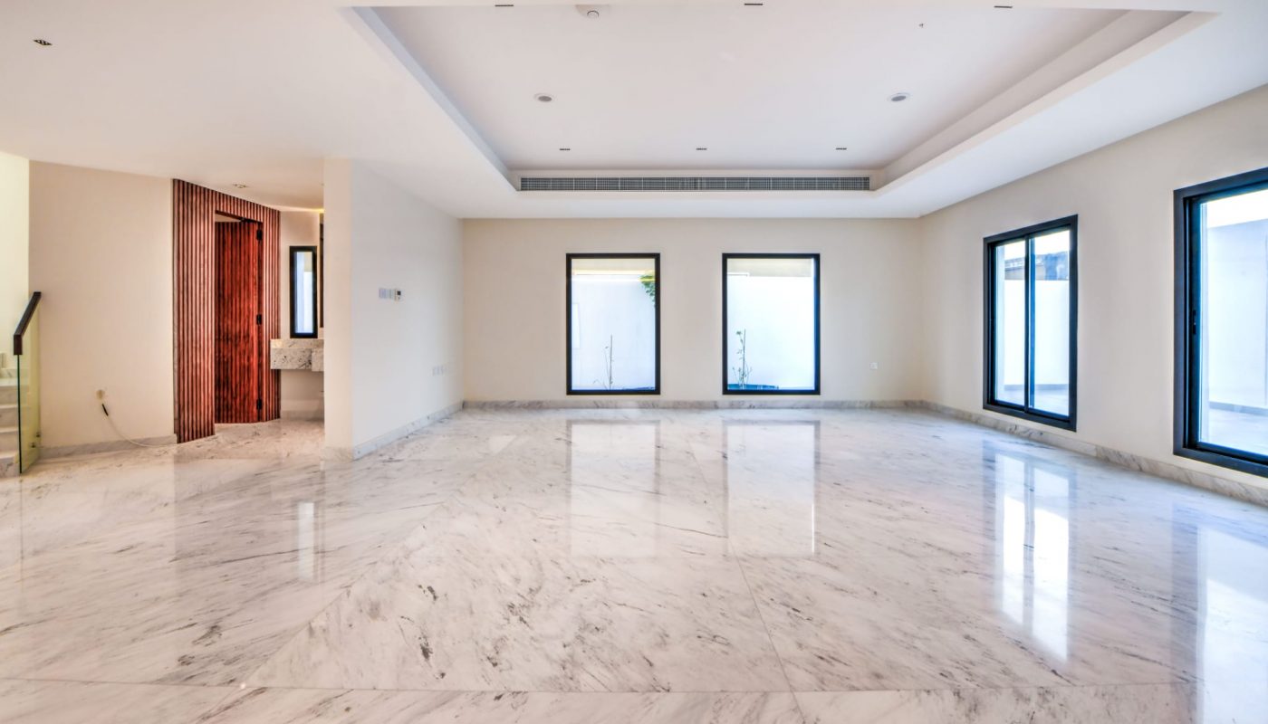 Spacious empty room with white marble floors, cream walls, recessed ceiling, three large windows, and a visible entryway to a bathroom.