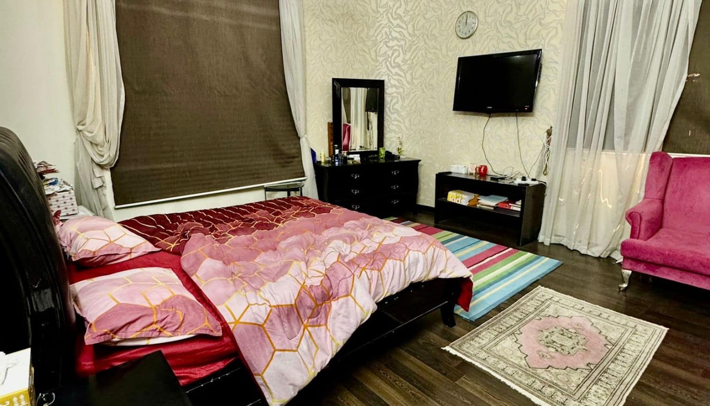 A bedroom with a double bed, patterned bedspread, dark wooden floor, dresser with mirror, wall-mounted TV, and a pink armchair. Ceiling fan and large windows with curtains are visible.
