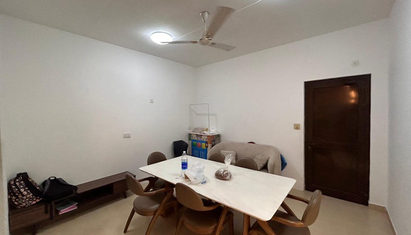 A dining room with a rectangular table, six chairs, a ceiling fan, a bench with bags, a water bottle on the table, and a closed door in the background.