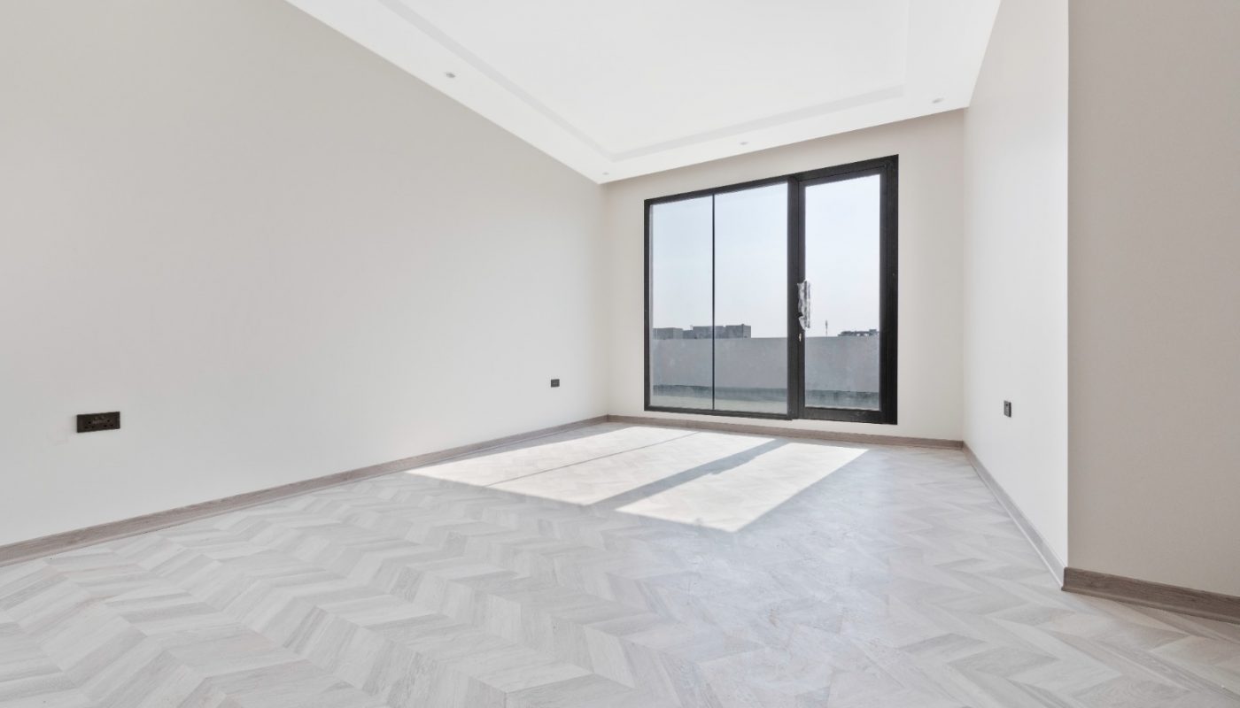 Empty room with light-colored herringbone flooring, white walls, recessed ceiling, and large sliding glass doors opening to a balcony with a city view.