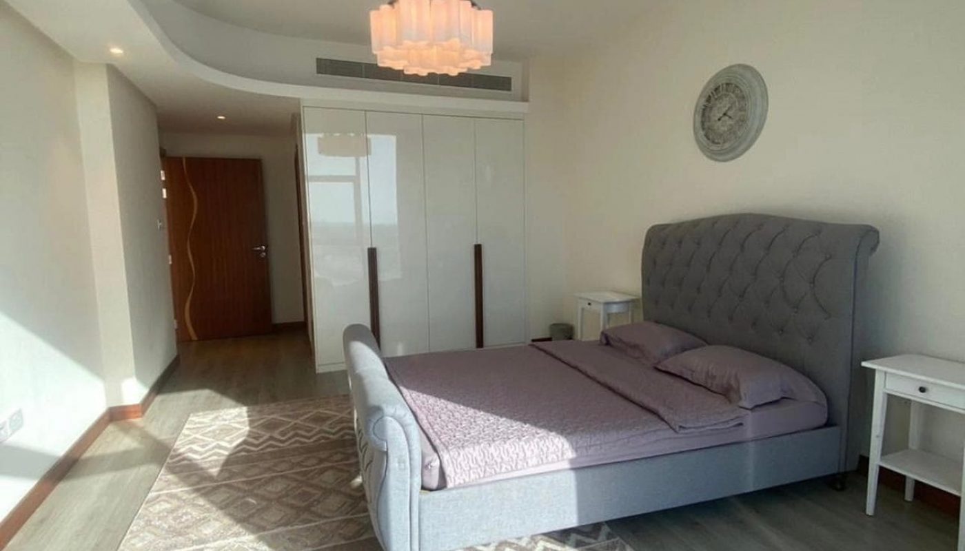 A modern bedroom with a grey upholstered bed, two white nightstands, a large wardrobe, a round wall clock, and a patterned rug on a wooden floor.