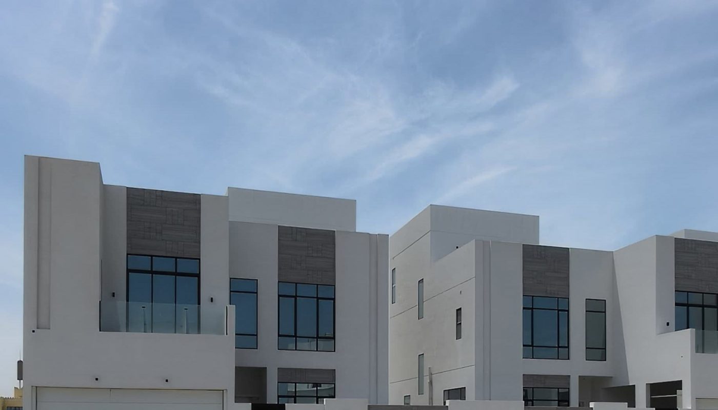 Two modern white two-story houses with large windows, separate garages, and a shared boundary wall under a partly cloudy blue sky.