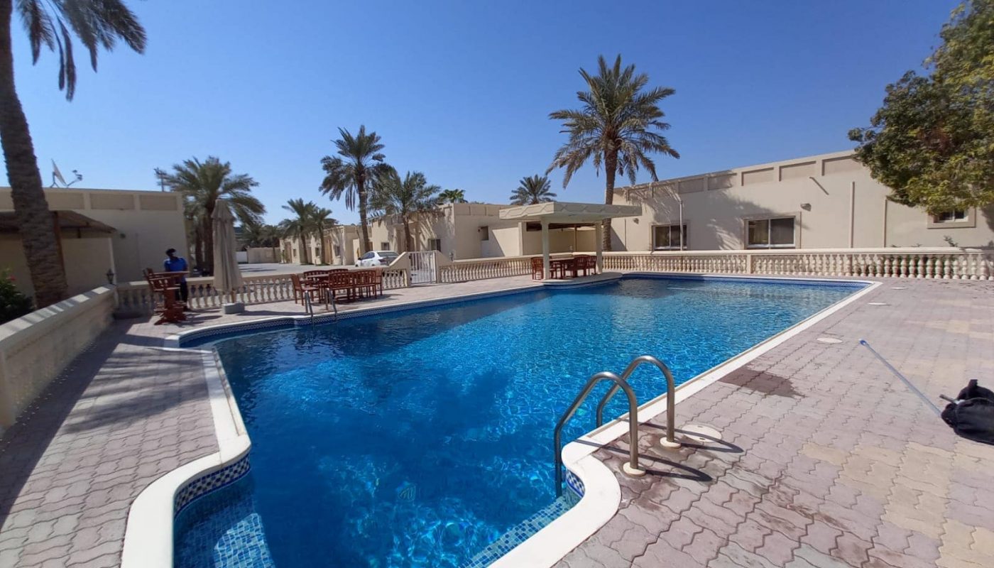 Outdoor swimming pool surrounded by palm trees and low buildings under a clear blue sky.