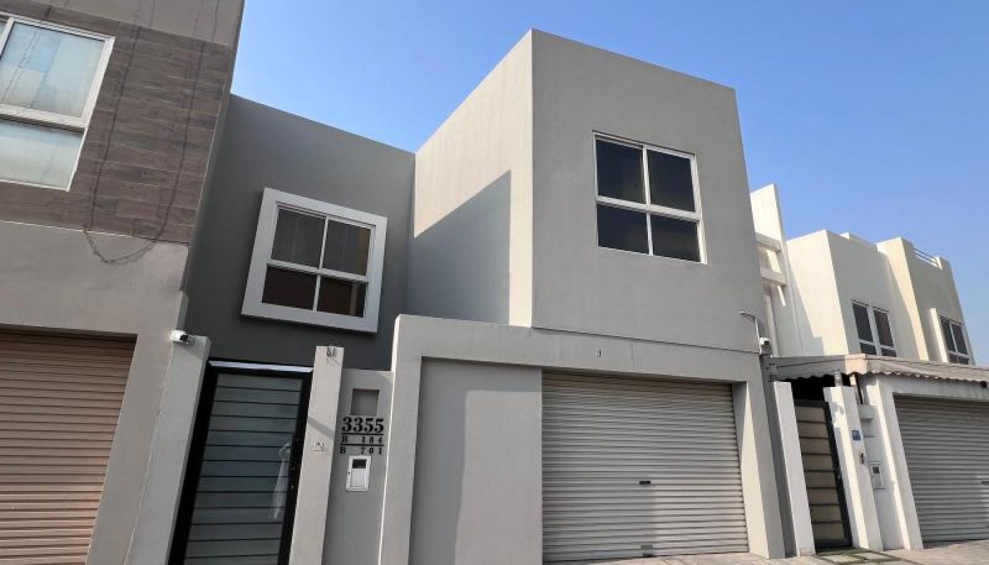 Modern two-story townhouse with gray exterior, two garages, large windows, and a gated entrance, under a clear blue sky.