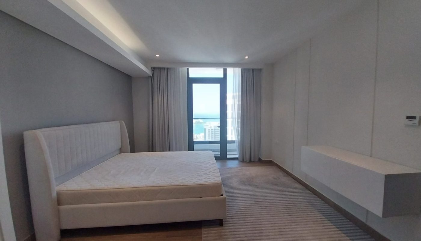 Minimalist bedroom with a white bed frame, mattress, wall-mounted storage, large window, light wood floor, and city view in the background.