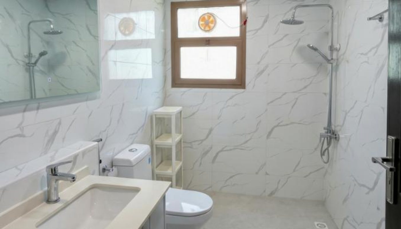 A modern bathroom with white marble walls, a wall-mounted shower, a toilet, a large mirror, a sink with a cabinet, and a window allowing natural light.