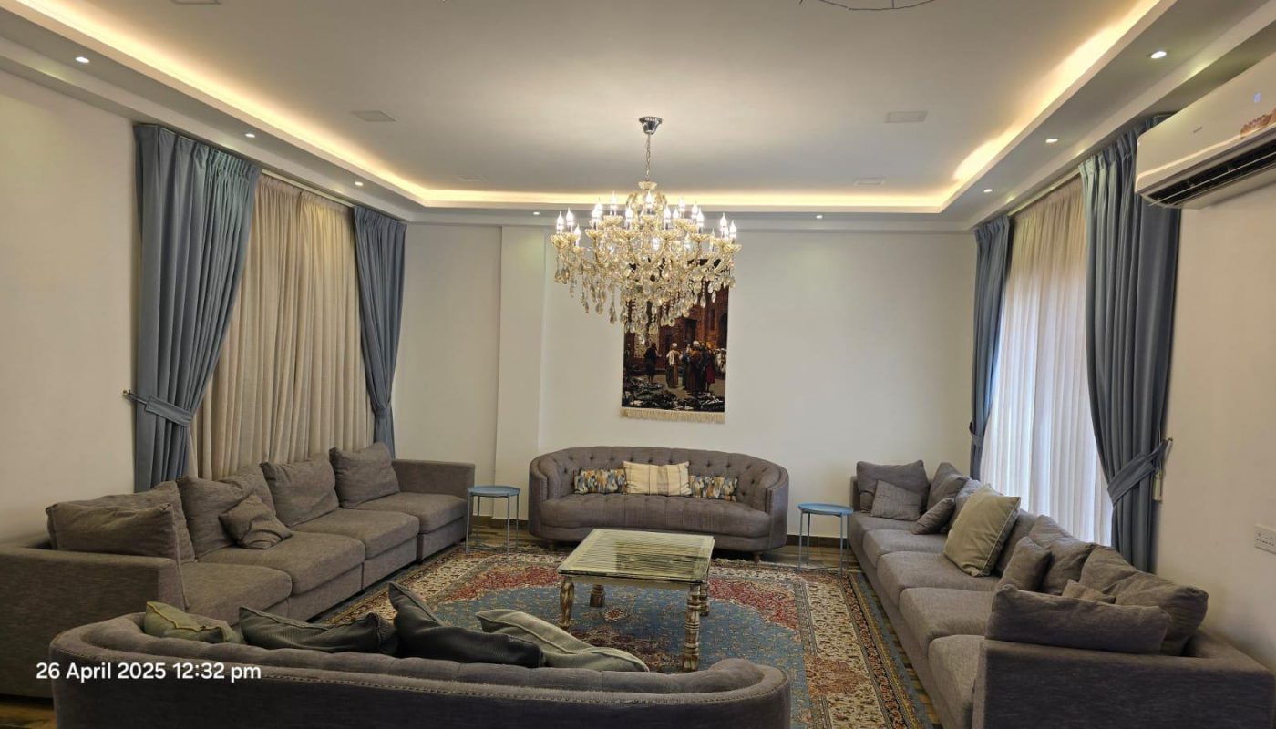 A living room with grey sofas arranged in a U-shape, a glass coffee table, a chandelier, blue curtains, a wall painting, and an oriental rug.