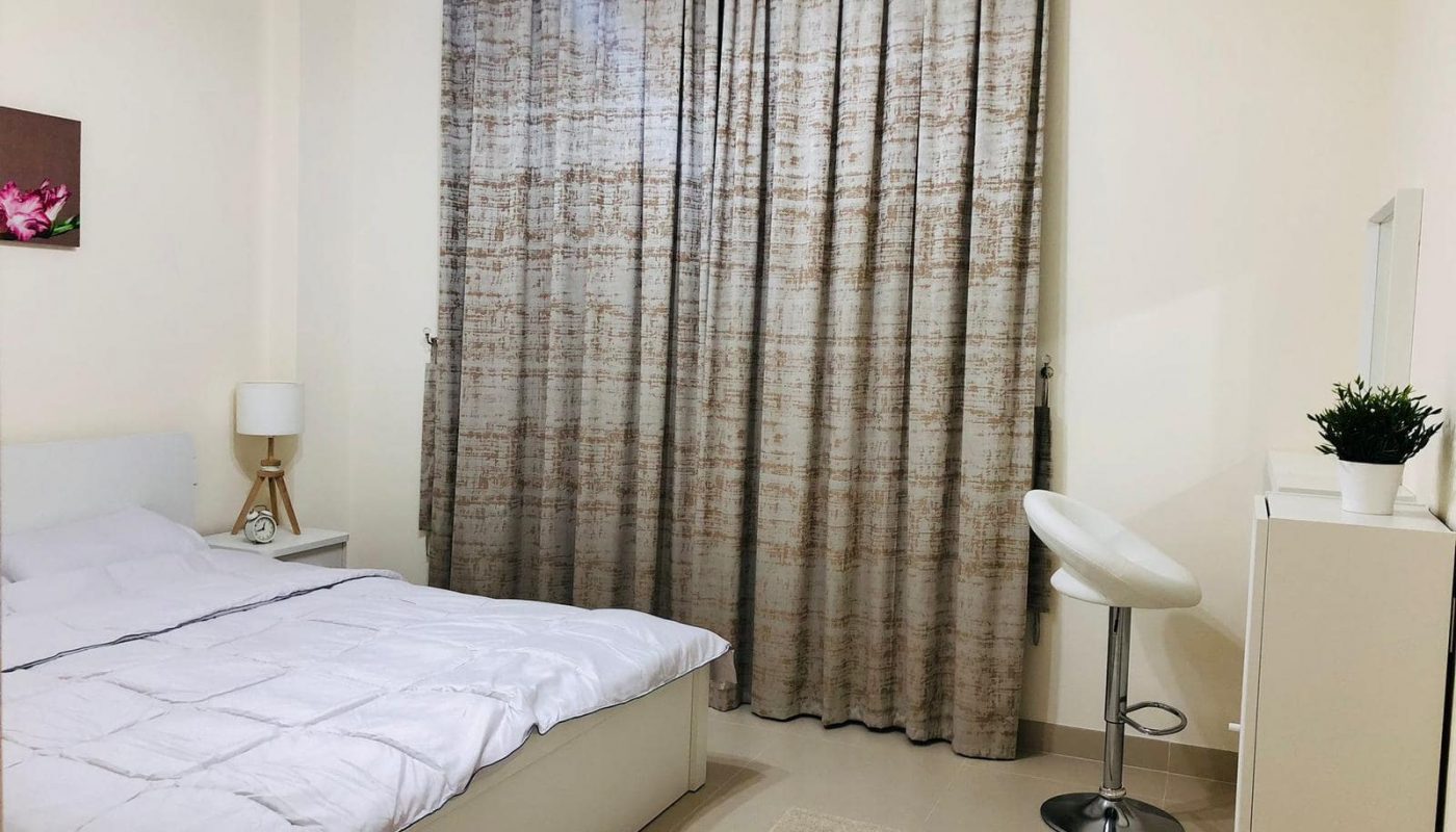 A neatly arranged 1bedroom flat in Marassi Boulevard features a cozy bedroom with a bed, side table, lamp, tall curtains, bar stool, vanity with mirror, and a potted plant. Light-colored walls and flooring add to the room's serene ambiance. For sale now!
