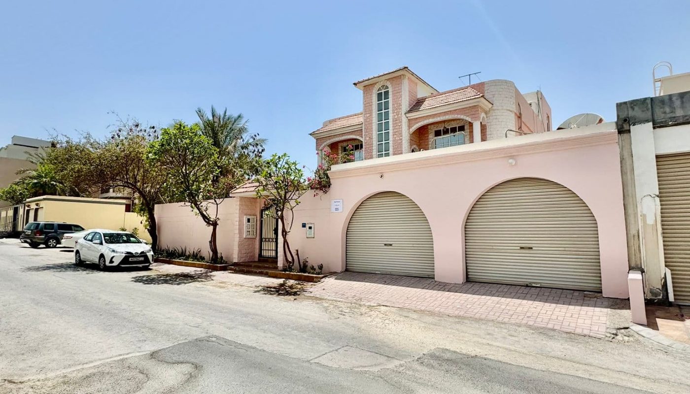 A residential street with a two-story pink house, three garage doors, parked cars, trees, and a clear blue sky.