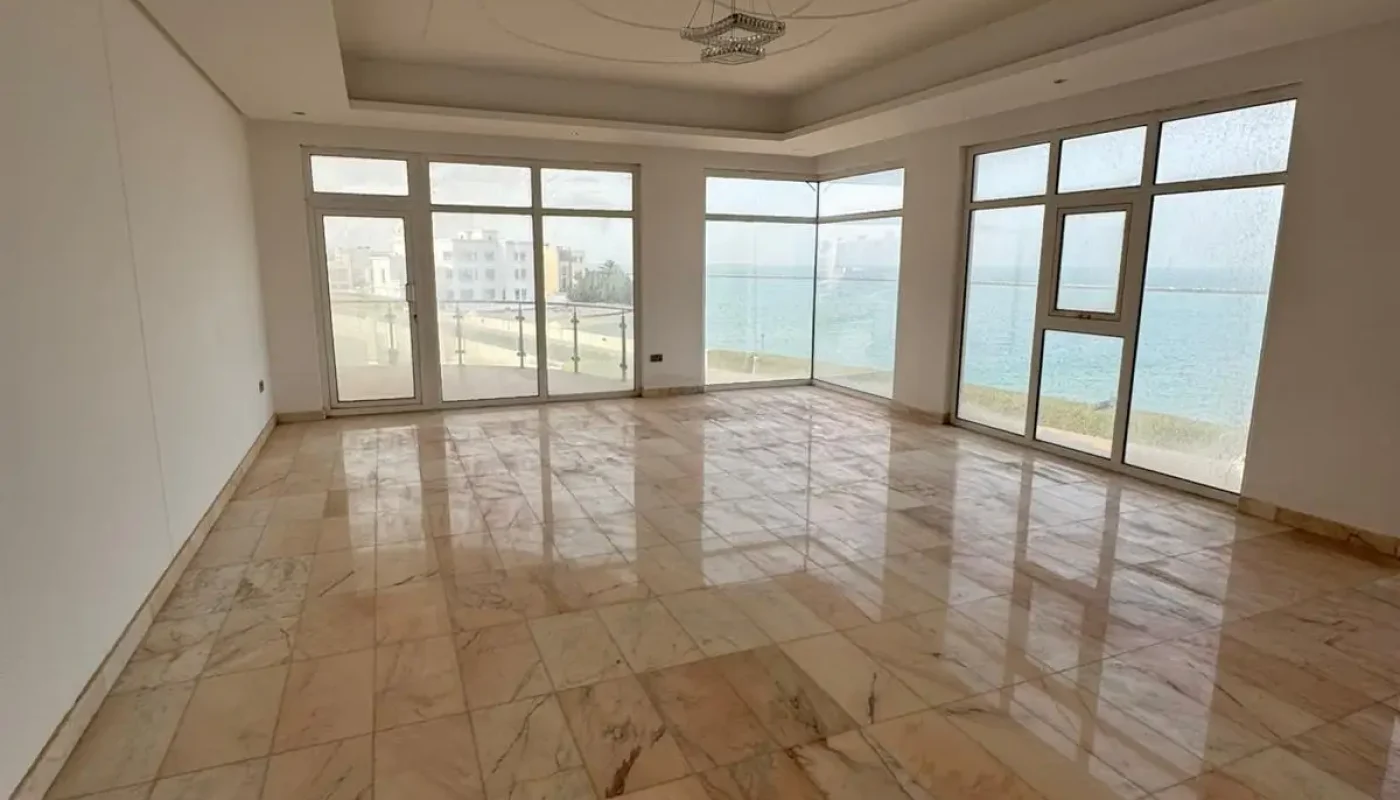 Spacious empty room with marble tile flooring, large windows, and a view of the water and nearby buildings outside.
