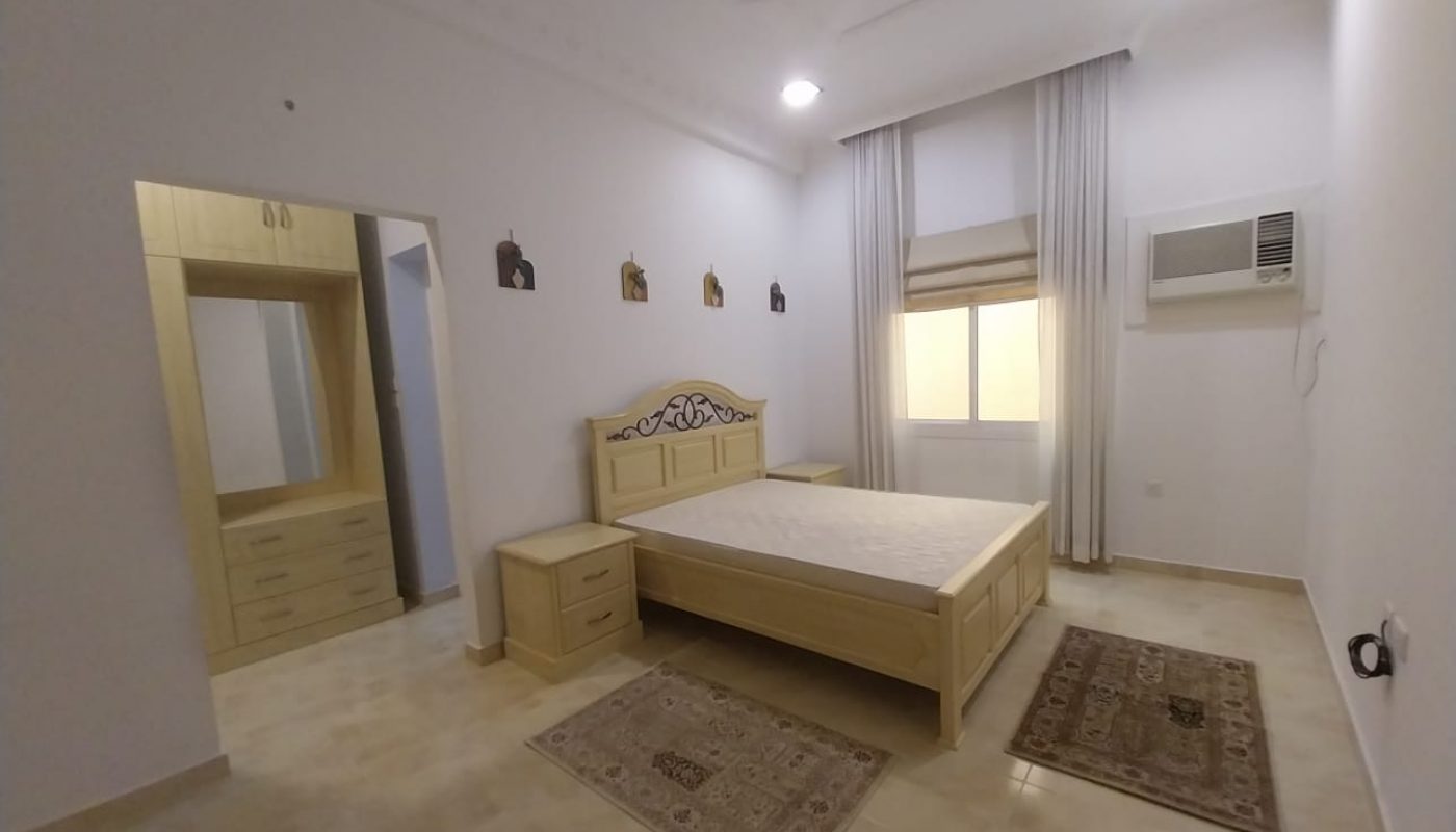 A simple bedroom with a wooden bed, nightstands, two rugs, a dressing table, wall-mounted art, window with curtains, and an air conditioner.