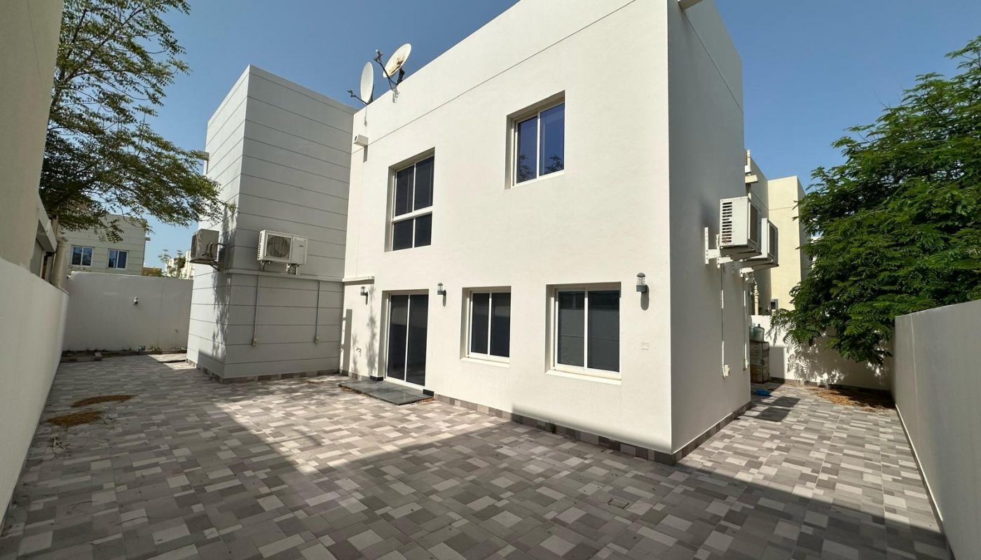 Two-story white modern building with several windows, air conditioning units, satellite dishes, and a spacious tiled courtyard under a clear sky.