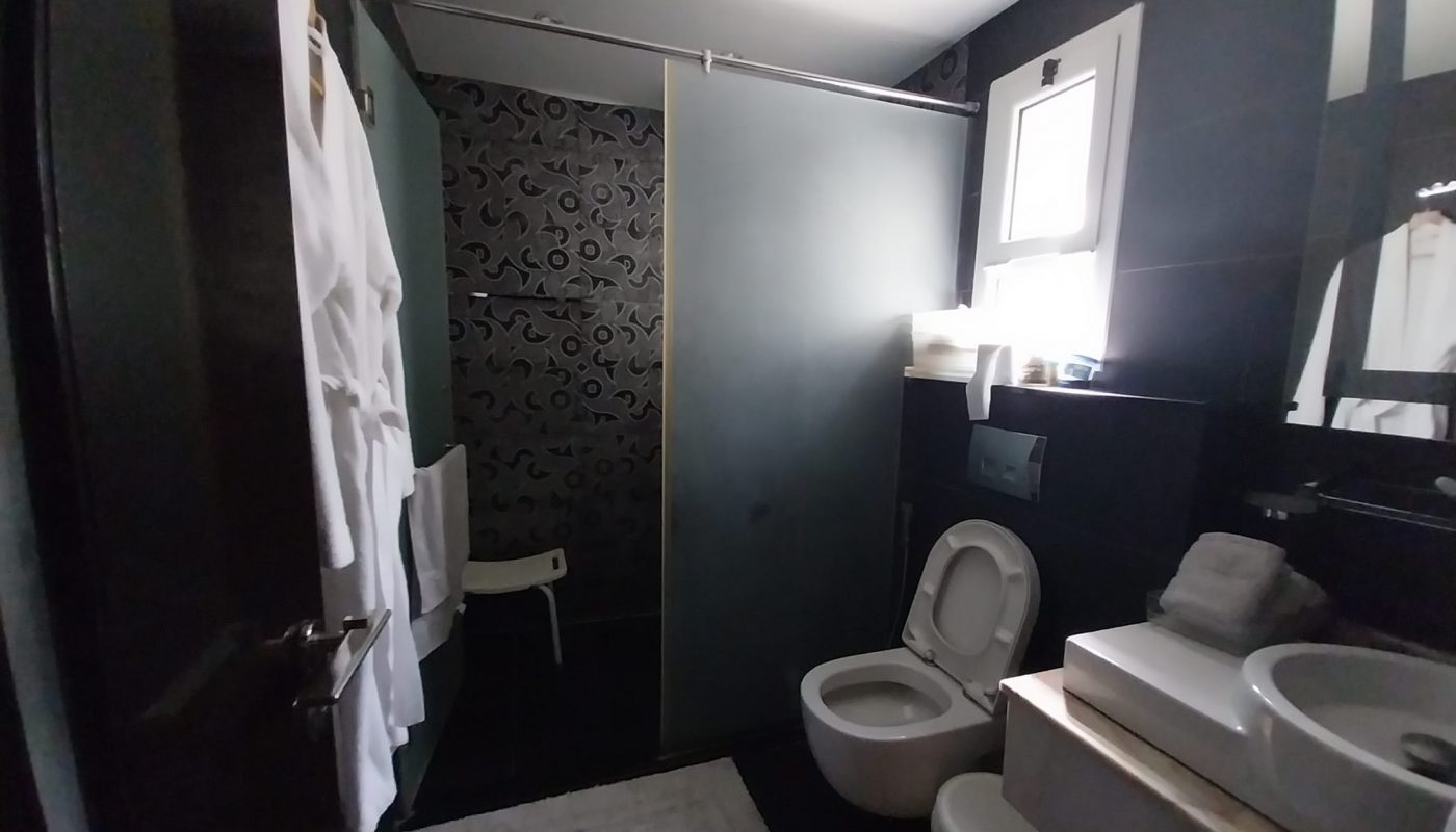 Modern bathroom with a toilet, bidet, sink, shower with glass door, white bathrobes on the door, and a window providing natural light.