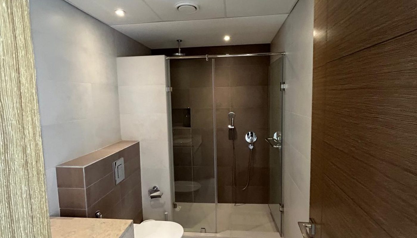Modern bathroom with a glass-enclosed shower, white toilet, wall-mounted flush, beige floor tiles, and wood accents on the door and cabinets.