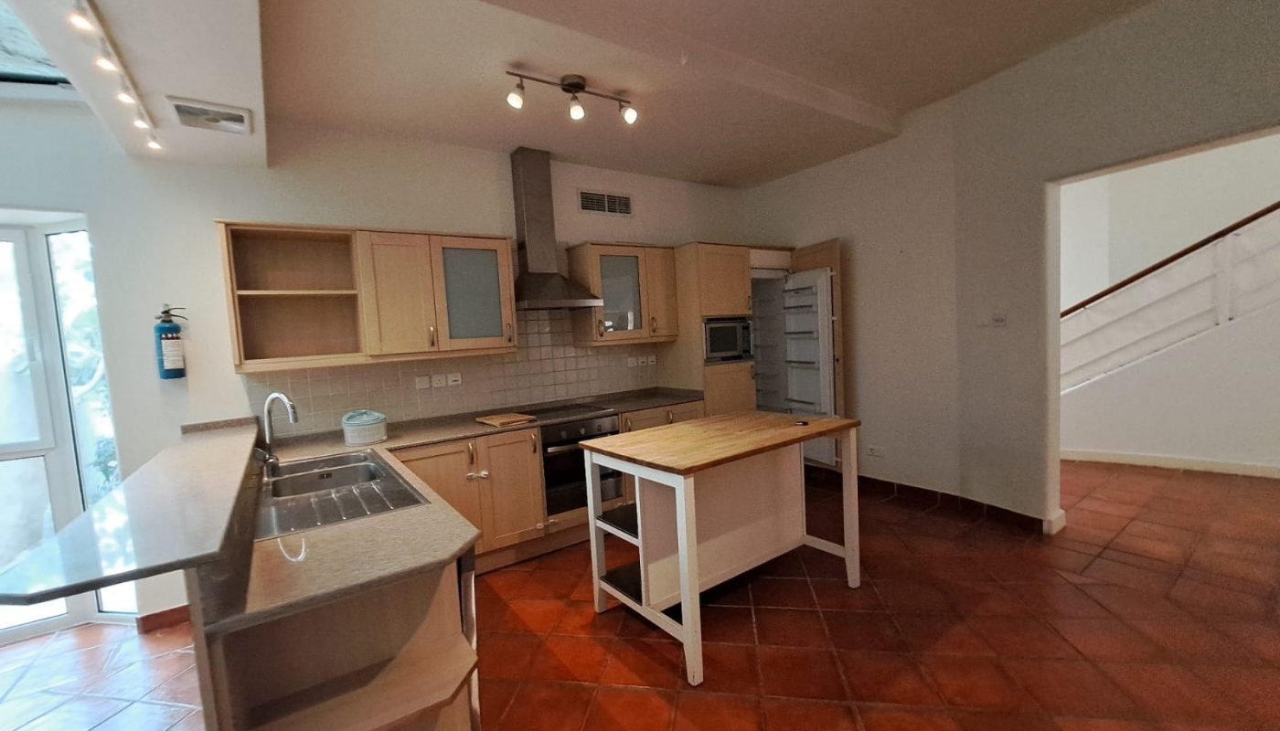 Spacious kitchen with wooden cabinets, center island, gas stove, stainless steel hood, open fridge, and red tile floor; a staircase is visible through an open doorway.