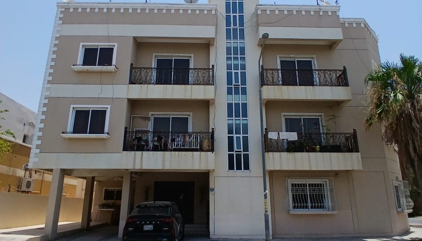 A three-story beige apartment building with balconies, a central glass stairwell, and a black car parked in front on a sunny day.