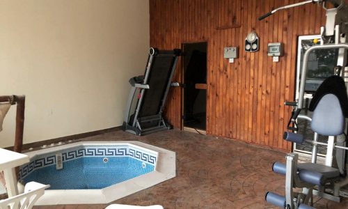 A small indoor gym with exercise equipment, a treadmill, and a corner hot tub surrounded by a tiled floor and wooden wall panels. A white plastic chair is in the foreground.