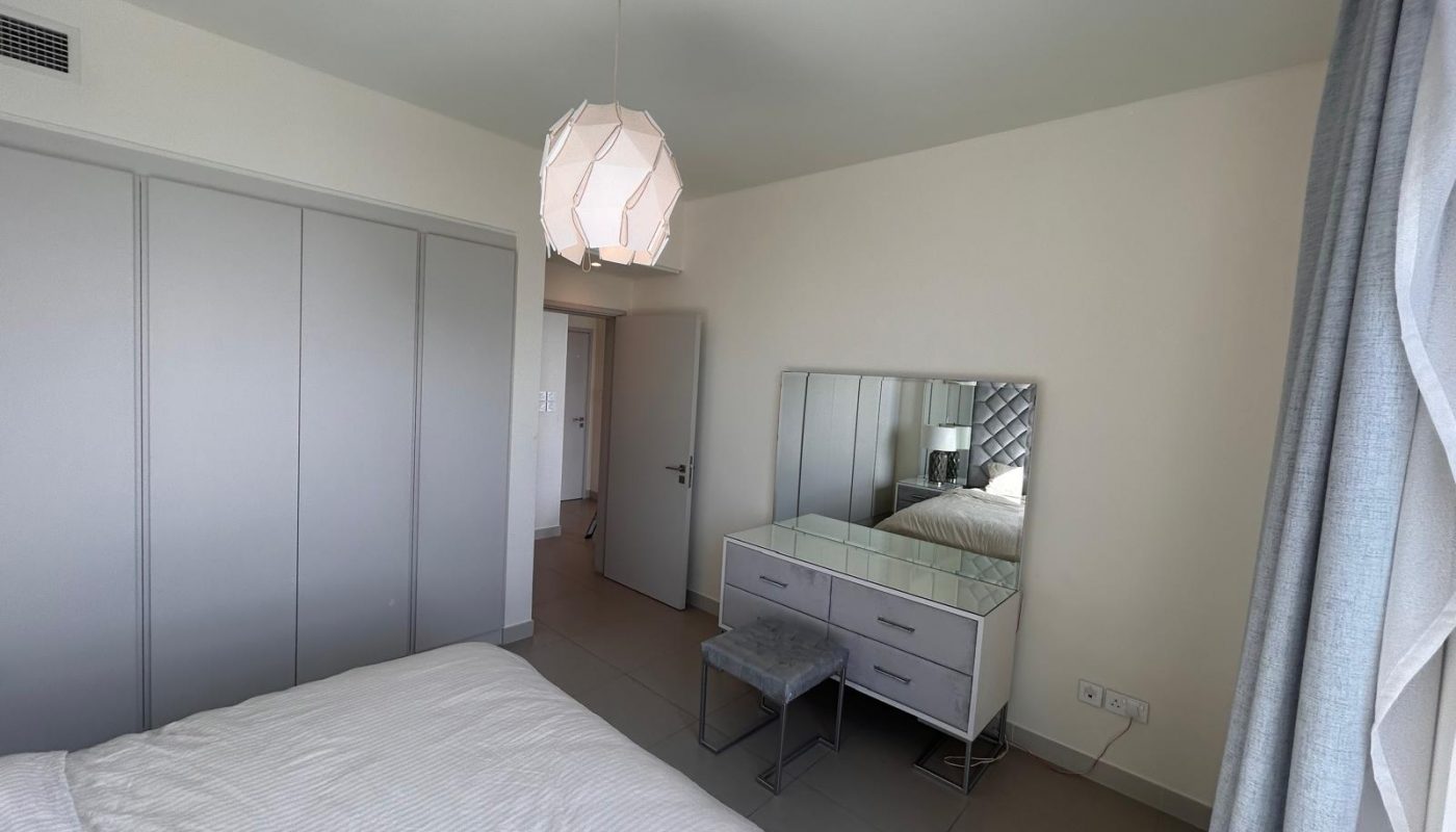 A bedroom with a white bed, gray wardrobe, mirrored vanity with a stool, and a geometric ceiling light. Light enters through a window with sheer curtains.
