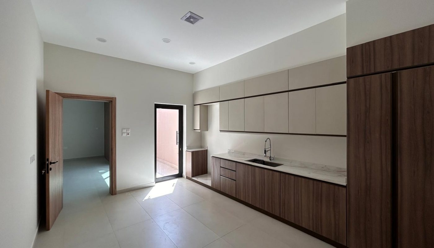 Modern kitchen with wooden cabinets, marble countertop, built-in appliances, sink, and a door leading to another room. Natural light enters through a glass door.