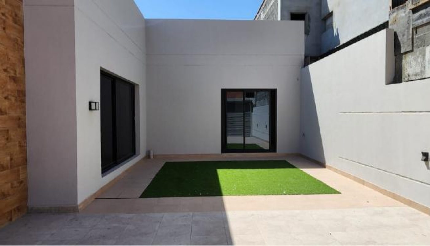 A modern, minimal courtyard with white walls, a sliding glass door, and a small rectangular patch of artificial grass in the center.