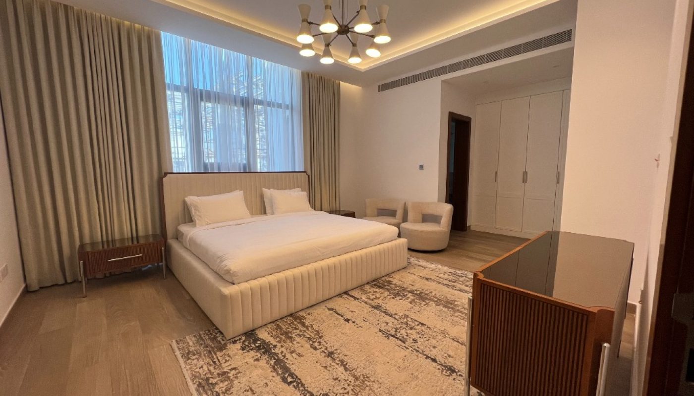 Modern bedroom with a large bed, two white chairs, a rug, a chandelier, floor-to-ceiling curtains, and built-in white wardrobes.
