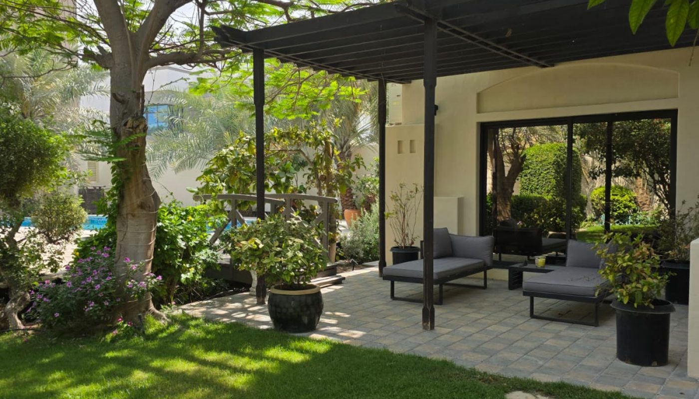 A patio with outdoor seating under a pergola, surrounded by lush green plants and trees, adjacent to a house with glass doors and a well-kept lawn.