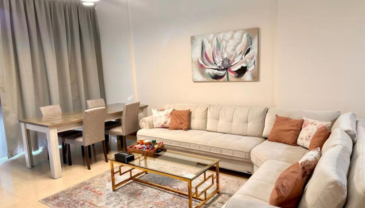 A living room with a white sectional sofa, glass coffee table, dining table with four chairs, floral wall art, and grey curtains.