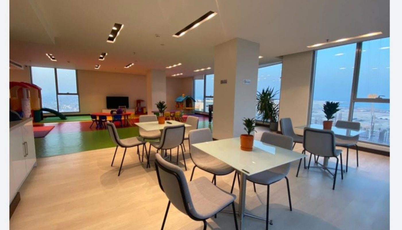 Bright, modern room with tables and chairs, potted plants, large windows, and a children's play area with toys and colorful mats in the background.