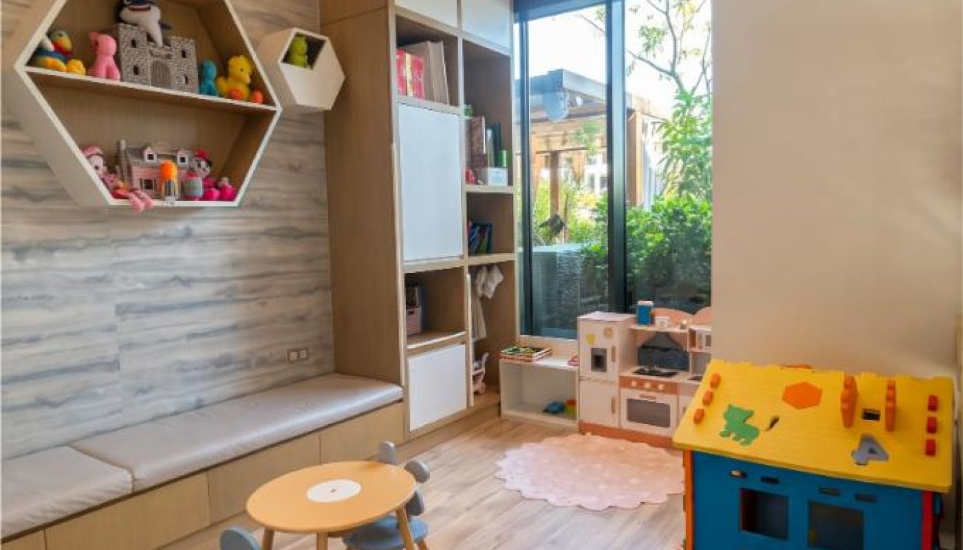 Modern playroom with wooden floor, shelves with toys, a play kitchen, a small table with chairs, a foam play mat, and a window showing greenery outside.