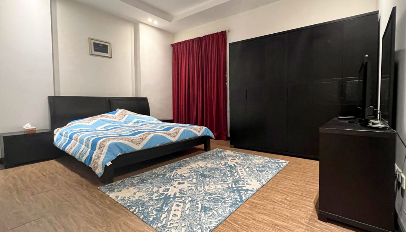 A modern bedroom with a double bed, blue-patterned rug, black wardrobe, TV on a stand, wooden floor, and red curtains covering the window.