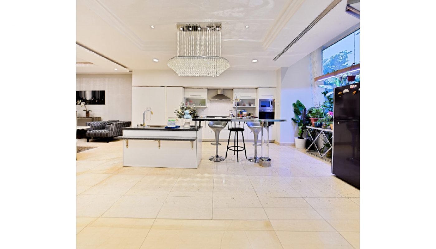 Modern kitchen with white cabinets, island counter with bar stools, large chandelier, tile flooring, and a window with plants.