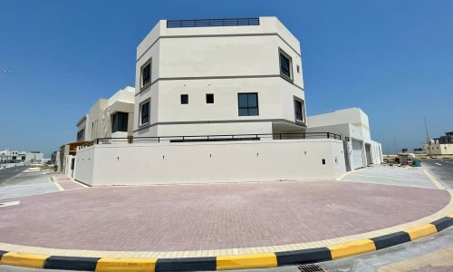 An elegant villa in Diyar Al Muharraq, this modern two-story beige building with black-trimmed windows sits on a corner lot with a circular driveway, surrounded by a sidewalk and yellow-black curbs under a clear blue sky. Now for sale!