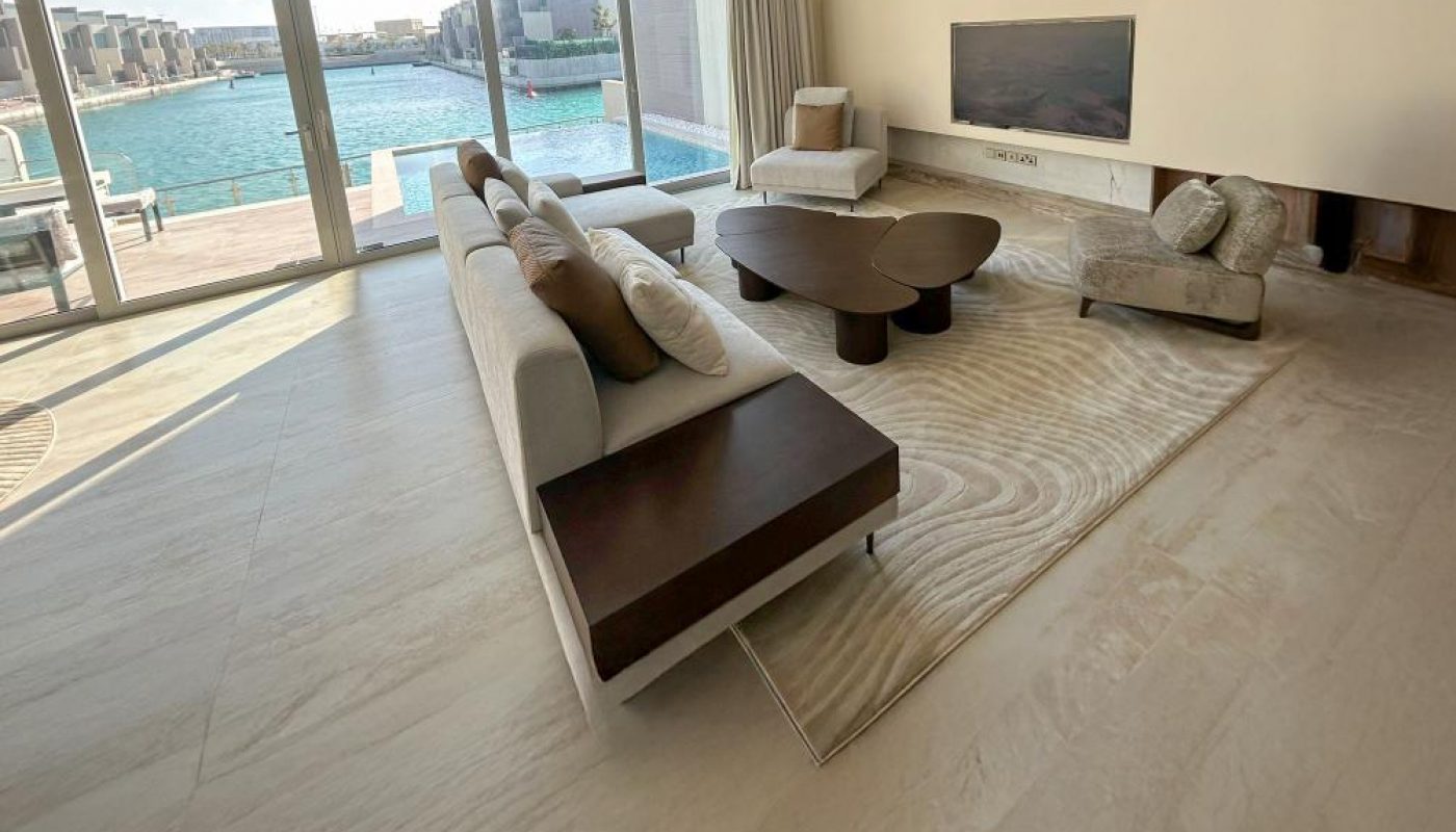 Modern living room with light tiled floor, beige sectional sofa, round coffee table, wall-mounted TV, and large windows overlooking a waterfront view.