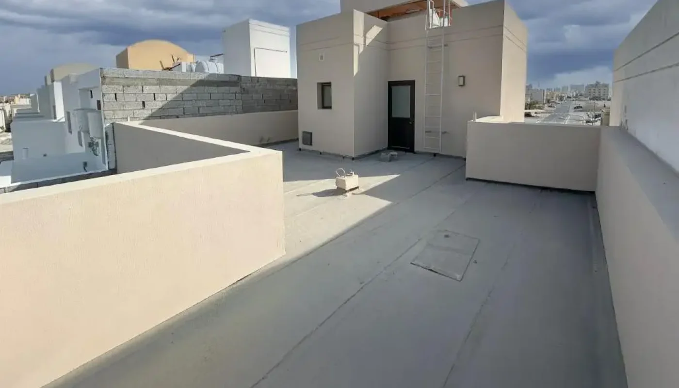 A flat rooftop with beige walls and minimal structures under a cloudy sky, featuring a small building and some equipment on the roof.