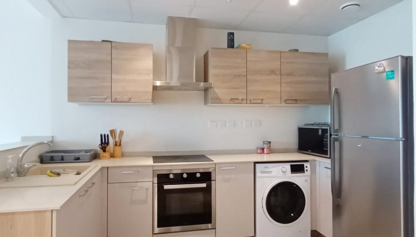 Modern kitchen with light wood cabinets, built-in oven, washing machine, stainless steel refrigerator, countertop appliances, and utensils, with a tiled floor and white ceiling.