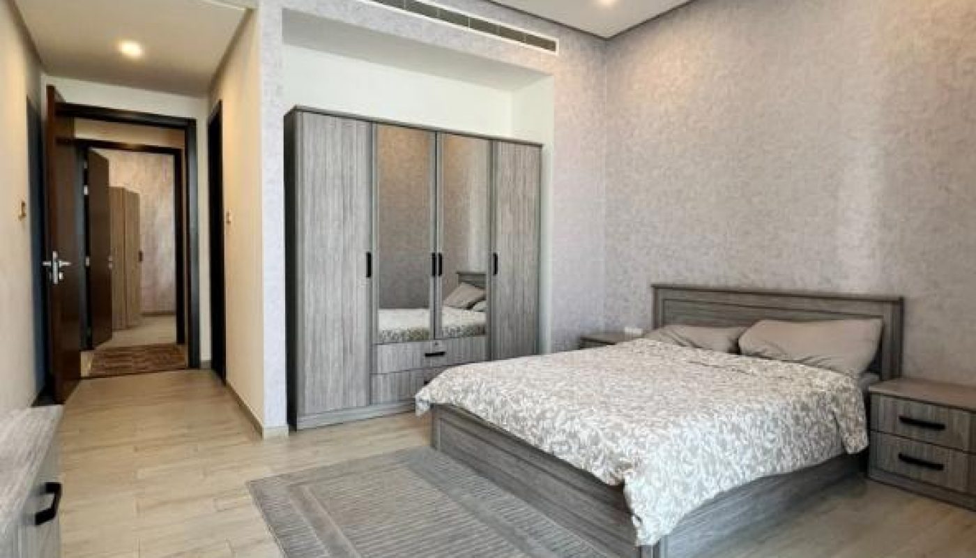 A modern bedroom with a double bed, bedside tables, a mirrored wardrobe, a dresser, and a gray rug on light wood flooring.
