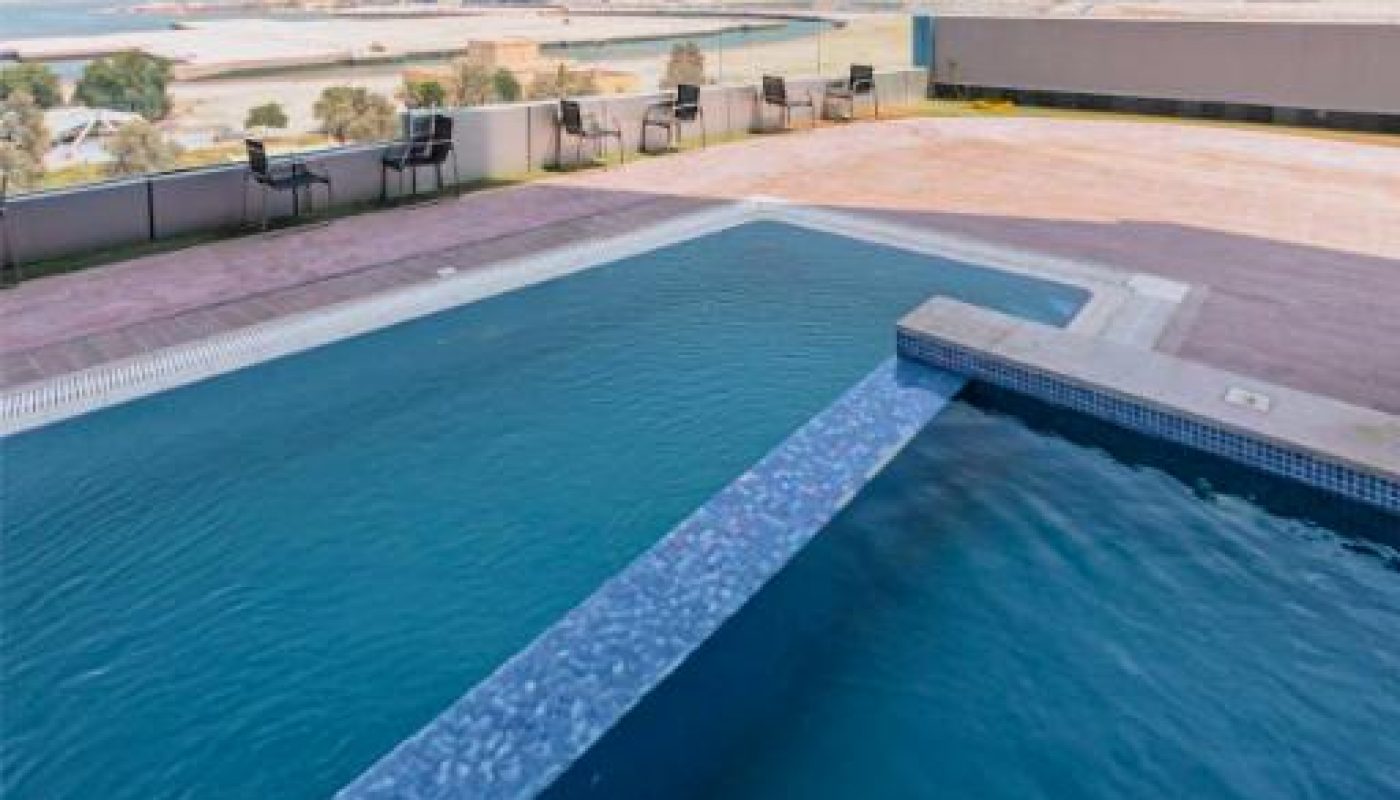 An outdoor swimming pool with a city skyline in the background; patio chairs are arranged along the pool's edge on a tiled deck under a clear sky.