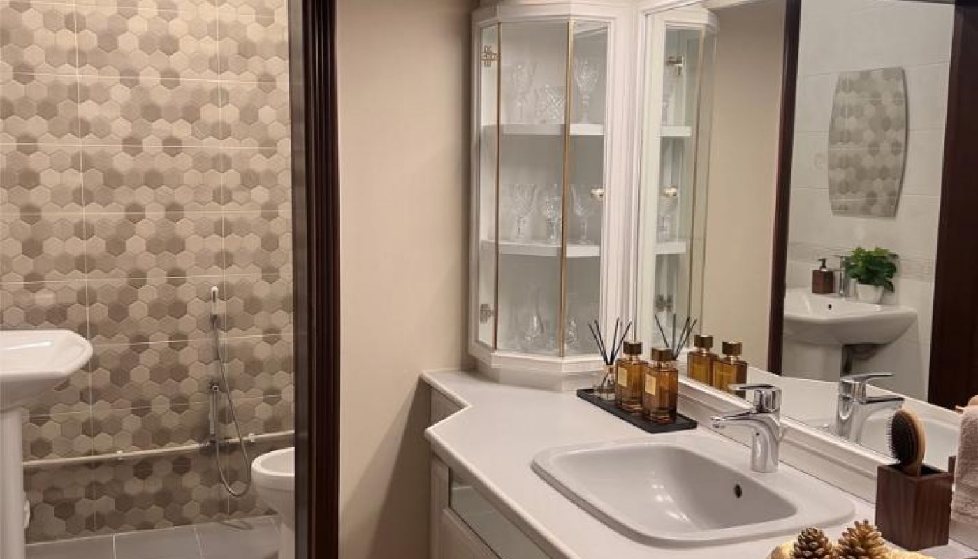 Modern bathroom with a lighted vanity, sink, and decorative items in the foreground; shower and toilet area with patterned tiles visible through a doorway.