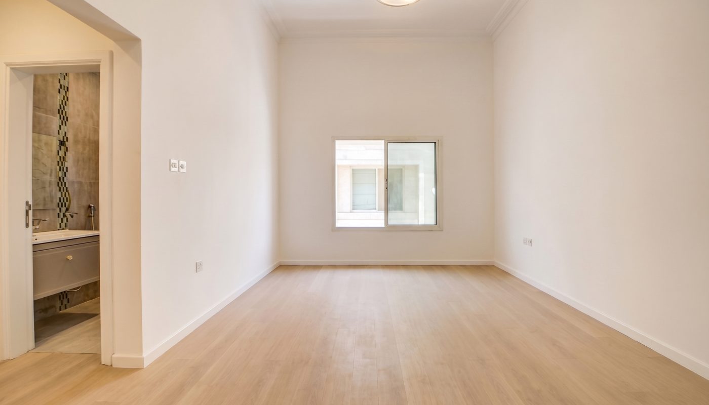 Empty room with light wood flooring, white walls, a ceiling light, a window, and an open doorway leading to a bathroom.