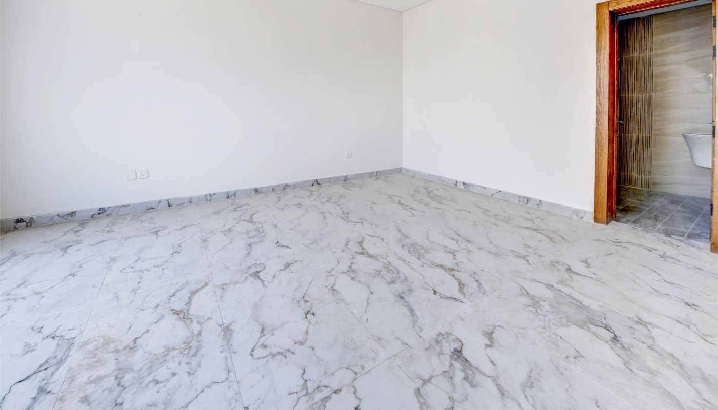 Empty room with white walls, recessed ceiling lights, marble-patterned tile floor, and an open wooden door leading to another tiled space.
