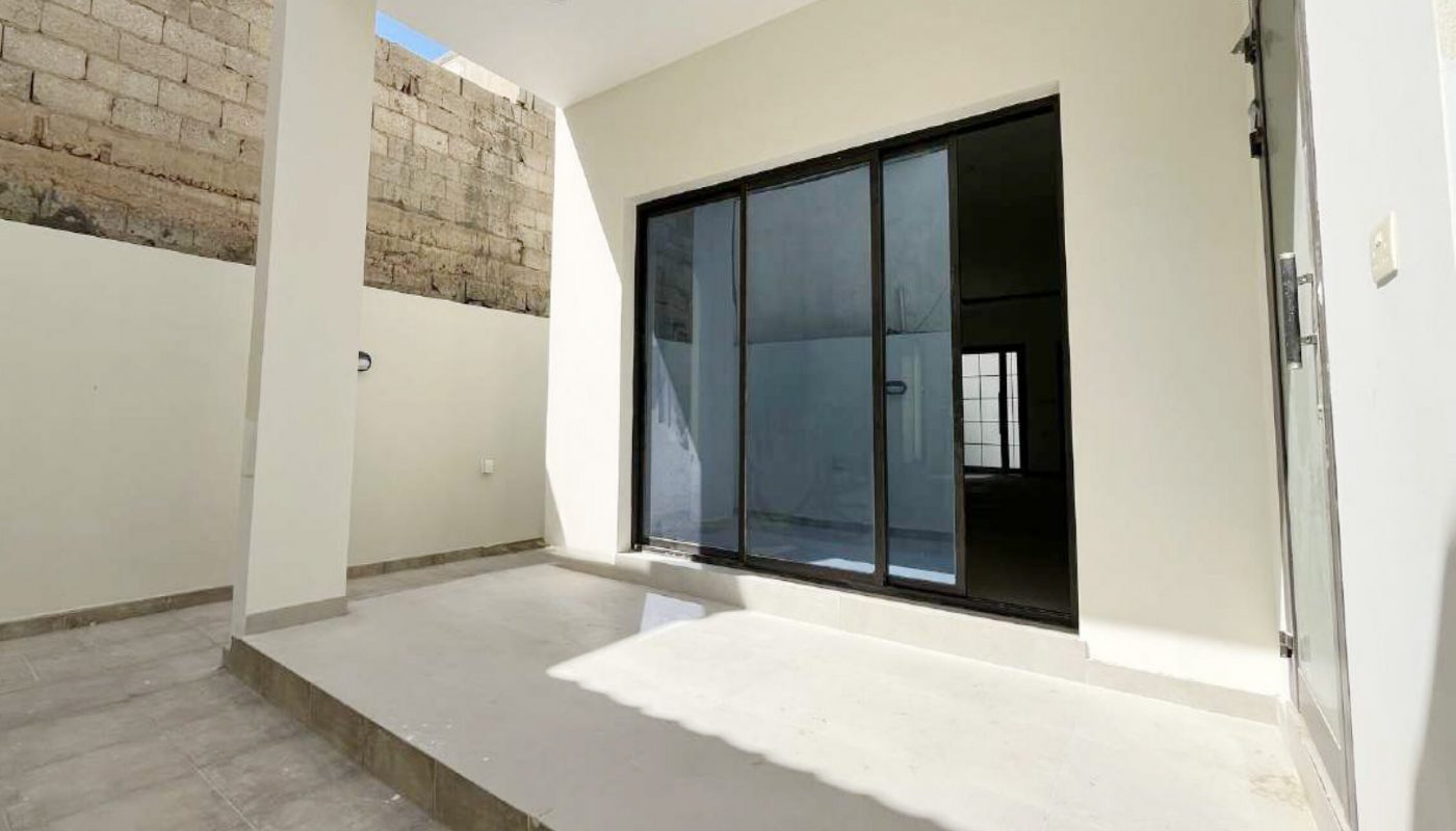 A modern, tiled patio with a small elevated platform, glass sliding doors, and beige walls, partially shaded by the building structure.