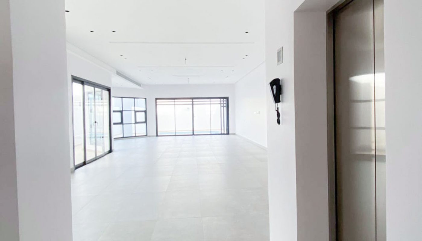 A bright, empty modern room with large windows, grey tiled floors, white walls, and an elevator door on the right side.